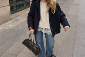 Look autunnale da donna con jeans blu dal taglio morbido, maglia panna con dettagli in pizzo, giacca corta blu navy e borsa scura, per un outfit casual chic dallo stile urbano.